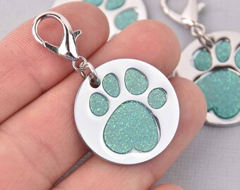 May include: A silver-colored, round pet tag with a glittery, light-green paw print design. The tag has a silver clasp for easy attachment to a collar. The paw print is a prominent feature, making it a stylish accessory for pets.
