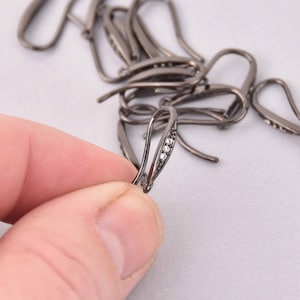 May include: A close-up of several gunmetal-colored earring hooks. One hook is held in a hand, showcasing three small, clear crystals. The hooks are designed for jewelry making and have a simple, elegant design.