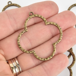 May include: Antique brass-colored clover-shaped charm with a textured surface. The charm has a small loop at the top for attaching to a chain or other jewelry component. The charm is held in a hand.