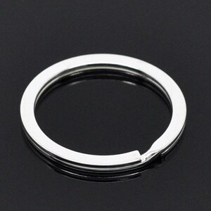 May include: A close-up shot of a silver-colored key ring. The key ring is circular with a split for adding keys. The key ring is made of a shiny, reflective metal and is set against a dark background.