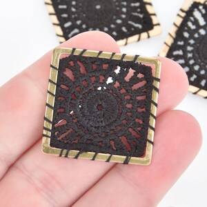 May include: A black and gold square-shaped decorative embellishment with a delicate lace pattern. The embellishment has a gold metal frame with a black trim.