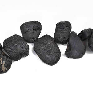 May include: A collection of dark, irregularly shaped stones with a rough, textured surface. The stones are a deep black color and appear to be of varying sizes. They are arranged against a plain white background.