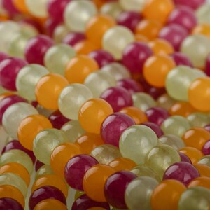 Puede incluir: Primer plano de hilos de cuentas de colores. Las cuentas son redondas y tienen un acabado pulido. Los colores incluyen tonos de naranja, rojo-p&uacute;rpura y verde p&aacute;lido. Las cuentas est&aacute;n ensartadas, creando un patr&oacute;n texturizado y visualmente atractivo.