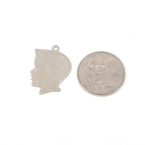 May include: Silver charm in the shape of a person's head, shown next to a 25 pence coin for size comparison.
