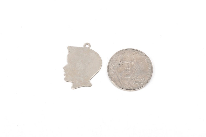 May include: Silver charm in the shape of a person's head, shown next to a 25 pence coin for size comparison.
