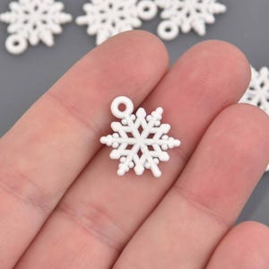 Puede incluir: Primer plano de un dije de copo de nieve blanco con un lazo en la parte superior para colgar. El copo de nieve tiene seis puntas simétricas con pequeños detalles redondos. Otros copos de nieve son visibles en el fondo.