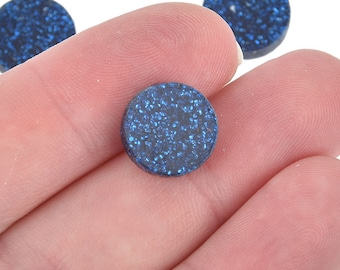 May include: Close-up of several small, round, dark blue glittered objects. One is held in a hand, with others visible in the background. The objects have a sparkly, textured appearance, with the glitter catching the light.