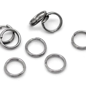 May include: A collection of small, silver-colored split rings, also known as key rings, scattered on a white surface. The rings are circular with a split for attaching items. The metal has a shiny, reflective quality.