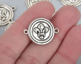 May include: Silver-toned metal connector with a fleur-de-lis design. The round charm has a raised border and two small loops for attachment. Several similar charms are visible in the background, suggesting a jewelry-making theme.