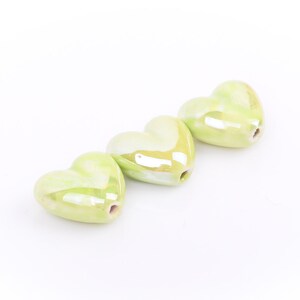 May include: Three light green heart-shaped beads with a glossy finish. Each bead has a hole for stringing. The beads are arranged diagonally on a white background. These beads are suitable for jewelry making.