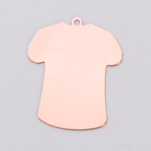May include: A copper-colored metal charm shaped like a t-shirt. The charm has a hole at the top for attaching to a necklace or bracelet.