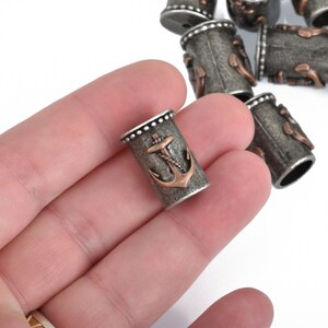 May include: A collection of cylindrical metal beads with an anchor design. The beads are primarily gray with bronze-colored anchor details. The anchor is a classic nautical symbol, and the beads have a rustic, antique appearance. These beads are suitable for jewelry making.
