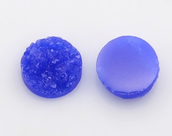 May include: Two round, blue, flat-backed cabochons. One cabochon has a smooth surface, while the other has a textured, crystal-like surface.