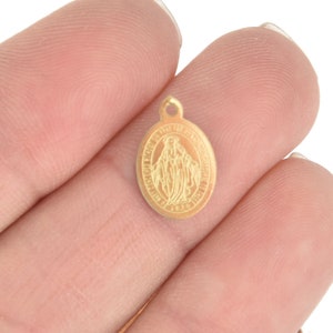 May include: A small, gold-colored oval pendant with a loop at the top. The pendant features an embossed image of a religious figure with text around the border. The pendant is held in a person's hand.