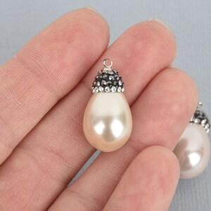 May include: A cream-colored, teardrop-shaped pendant with a silver-colored bail. The top of the pendant is adorned with small, dark gray and clear crystals. The pendant is held in a person's hand.