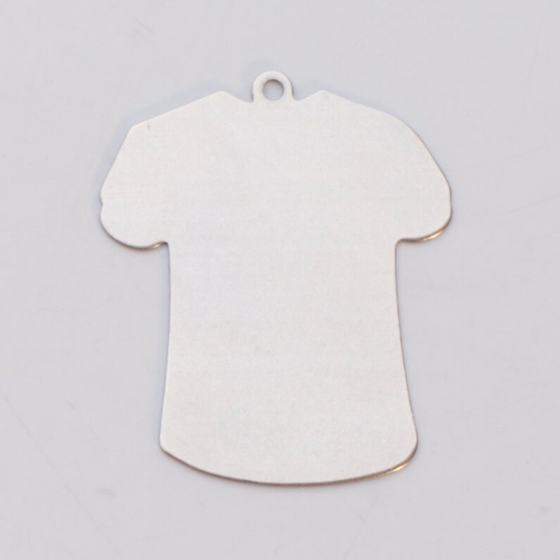 May include: A silver metal charm shaped like a t-shirt. The charm has a hole at the top for hanging.