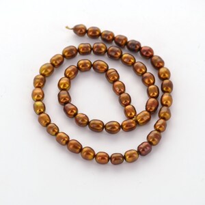 May include: A strand of lustrous, oval-shaped, bronze-colored pearls. The pearls have a warm, iridescent sheen, with hints of gold and reddish-brown. The pearls are strung together, forming a necklace or a similar jewelry piece.