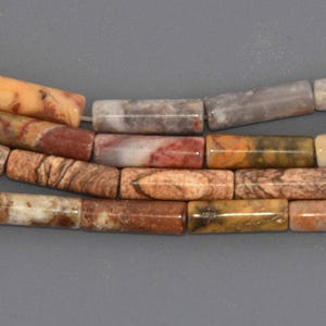 May include: Close-up of three strands of rectangular gemstone beads. The beads display a variety of colors, including shades of brown, red, yellow, and gray. The beads are strung together, showcasing their natural patterns and textures.
