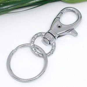 May include: A silver-colored metal swivel clasp with a key ring. The clasp has a spring-loaded lever for easy attachment. The key ring is a simple, circular design. The hardware is likely used for keychains, lanyards, or bag accessories.