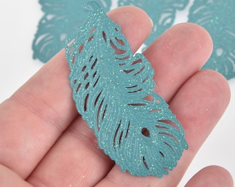 May include: Close-up of a teal-colored, glitter-covered feather-shaped craft embellishment. The feather has intricate cut-out details and is held in a hand. Several similar feathers are visible in the background.