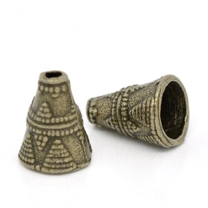 May include: Two antique bronze-colored cone-shaped beads with intricate geometric patterns. The beads have a wide opening at one end and a smaller hole at the other, suitable for stringing. The design includes triangles and dotted lines.