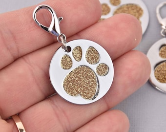 May include: A silver-colored, round pet tag with a gold glitter paw print design. The tag has a silver clasp for easy attachment to a collar or leash. Additional tags with paw prints are visible in the background.
