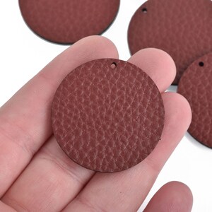 May include: A collection of burgundy-colored, round leather pieces with a textured surface. Each piece has a small hole at the top. The leather pieces are in various sizes and are suitable for crafting projects.