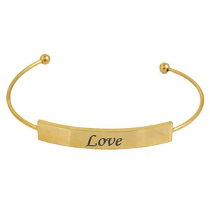 May include: A gold-colored cuff bracelet with the word "Love" engraved in black on a rectangular plate. The bracelet has a curved design with small ball accents at each end.