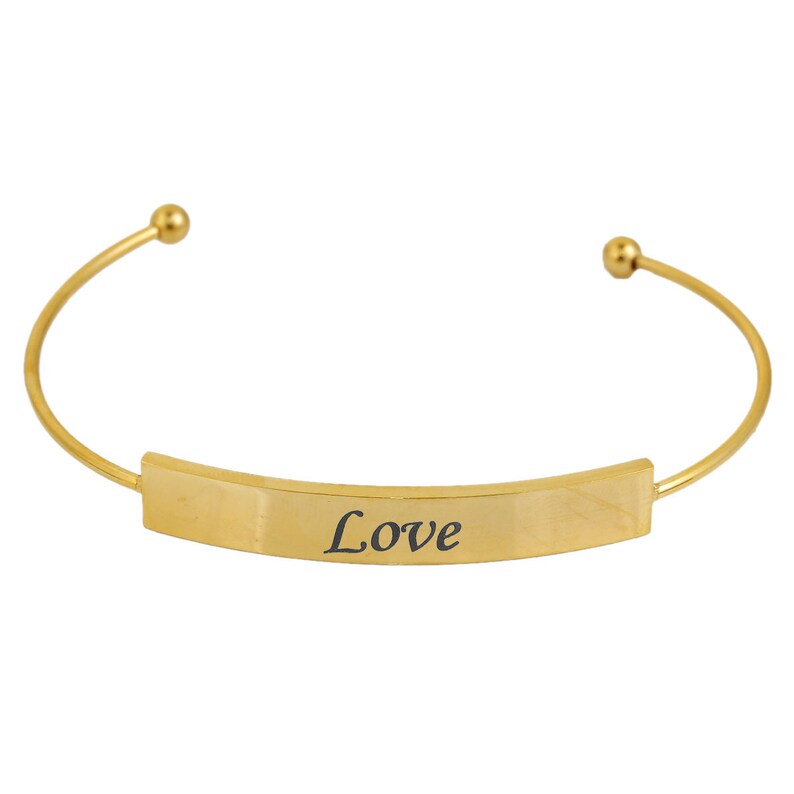 May include: A gold-colored cuff bracelet with the word "Love" engraved in black on a rectangular plate. The bracelet has a curved design with small ball accents at each end.