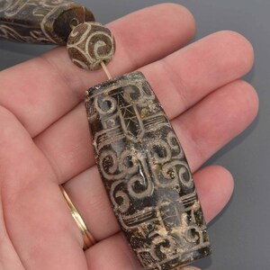 May include: Close-up of a strand of antique beads. The beads are a dark, mottled brown color with intricate carved designs. The designs include swirling patterns and geometric shapes. The beads are strung together with a thin, light-colored string.
