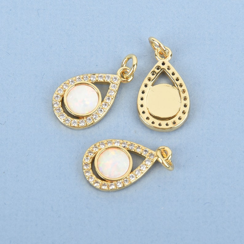 Puede incluir: Colgantes dorados en forma de l&aacute;grima con una piedra central de &oacute;palo blanco y un halo de peque&ntilde;os cristales transparentes. Los colgantes tienen un lazo en la parte superior para sujetarlos a una cadena u otras joyas. El fondo es azul claro.