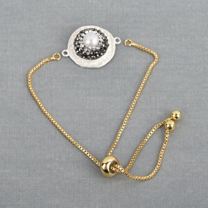 May include: A gold chain bracelet with a white and black round charm featuring a pearl and rhinestones. The bracelet has an adjustable sliding clasp.