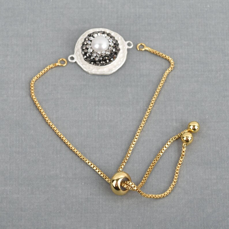 May include: A gold chain bracelet with a white and black round charm featuring a pearl and rhinestones. The bracelet has an adjustable sliding clasp.