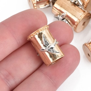 May include: A gold-colored metal bead with a silver-colored bird design. The bead is cylindrical with a decorative rim at the top. Several similar beads are visible in the background, suggesting a jewelry-making theme.