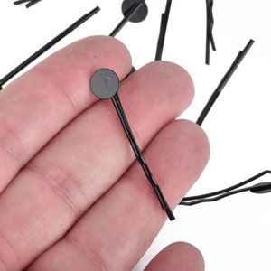 May include: A collection of black bobby pins with a small, round, flat head. The bobby pins are made of metal and have a simple, functional design. The pins are scattered on a white background.