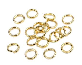 May include: A group of 15 gold jump rings, scattered on a white background. The jump rings are small and circular, with a slightly open gap on one side.
