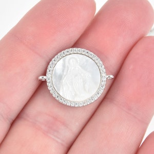 May include: Oval-shaped pendant with a white mother-of-pearl inlay featuring a religious figure. The pendant is surrounded by a border of clear cubic zirconia stones.