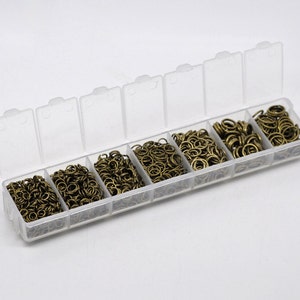 May include: A clear plastic storage box with seven compartments filled with gold jump rings. The box is open and the jump rings are visible.