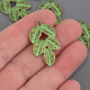 Op de afbeelding: Een groene en bruine bladbedel met een lus om op te hangen. De bedel heeft een lichtgroene emaille afwerking met bruine accenten op de bladeren. De bedel is waarschijnlijk voor het maken van sieraden of andere knutselwerkjes.