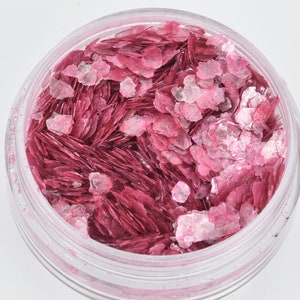 May include: A clear plastic jar filled with pink and white mica flakes. The flakes have a layered, textured appearance, with varying shades of pink and white. The jar is round and transparent, allowing the contents to be fully visible.