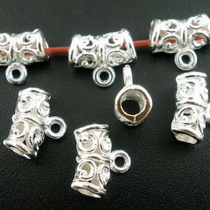 May include: Silver-toned metal jewelry findings with intricate scrollwork and a loop for attaching to other components. The findings are cylindrical with a decorative pattern and a small loop for attaching to other components.