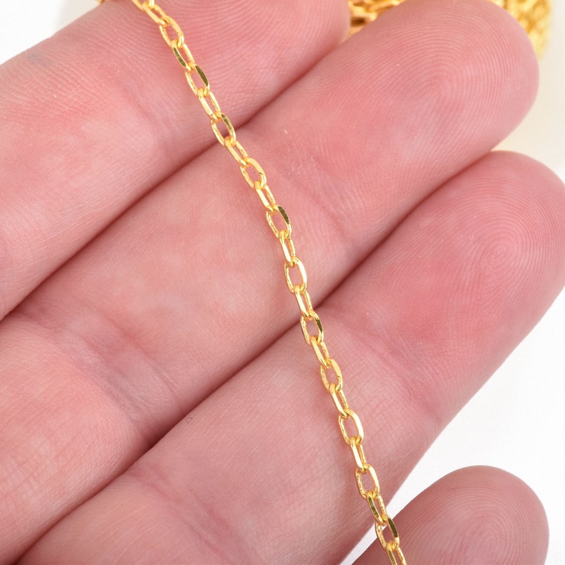 May include: Close-up of a delicate gold-colored chain with oval links. The chain is held in a hand against a white background. The chain is a bright, shiny gold color.