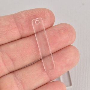 May include: A clear, rectangular acrylic charm with a small hole at the top for stringing. The charm is held in a hand, showcasing its transparent quality. This item is suitable for jewelry making or craft projects.