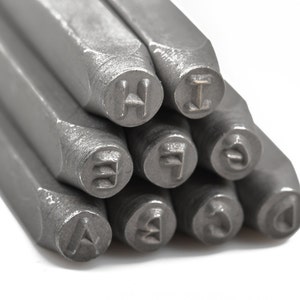 May include: Close-up of a collection of metal letter stamps. The stamps are arranged in a diagonal pattern, with the letters A, B, E, F, H, I, J, 3, and 4 visible. The stamps are made of a gray metal and are on a white background.