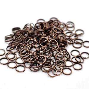 May include: A pile of small, open jump rings in a bronze color. These metal rings are round with a split, and are used for jewelry making and crafts. The rings are scattered on a white surface.