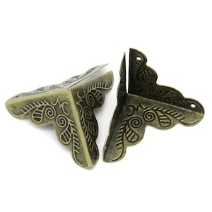 May include: Two antique brass-colored corner brackets with decorative floral and swirl patterns. The metal corner protectors are designed to be mounted on the corners of a box or other item to provide reinforcement and a vintage aesthetic.