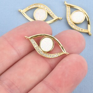 May include: Gold-colored eye-shaped charm with a white center and a border of small, clear crystals. The charm is held in a person's hand against a light blue background. The charm is approximately 1 inch long.