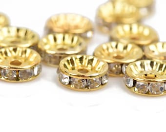 May include: Close-up of several gold-colored metal beads with a ring of clear, sparkling rhinestones around the center. The beads have a shallow, bowl-like shape with a central hole. The background is white.
