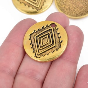 May include: Gold-colored metal pendant with a geometric diamond design. The pendant is round with a small hole at the top for stringing. The diamond design is framed by a decorative border. Other similar pendants are visible in the background.