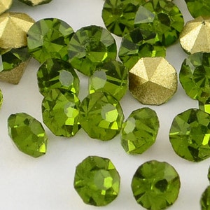 May include: Close-up of various green and gold faceted rhinestones. The green rhinestones are round and oval-shaped, while the gold ones have a geometric, multi-sided design. The gemstones are scattered on a white surface.